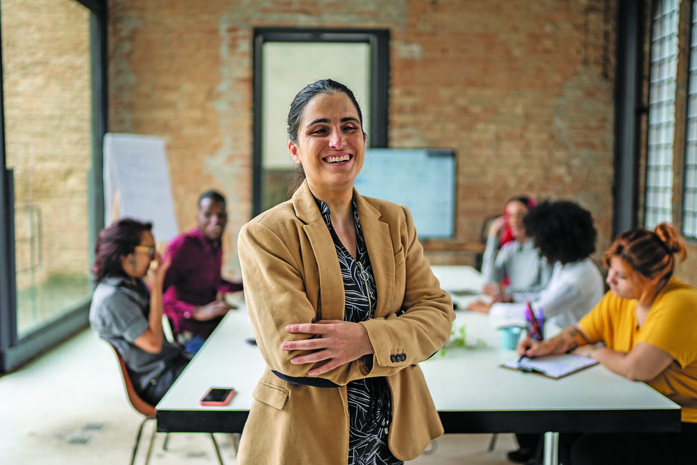 A women with sight loss smiling with her arms crossed. She is standing in front of a group of people sitting around a boardroom table