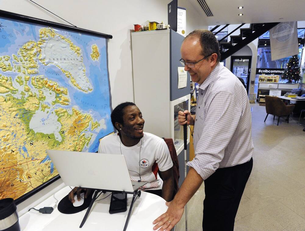 A young man sitting at a computer. Another man holding a white man stands beside him, engaging with the computer