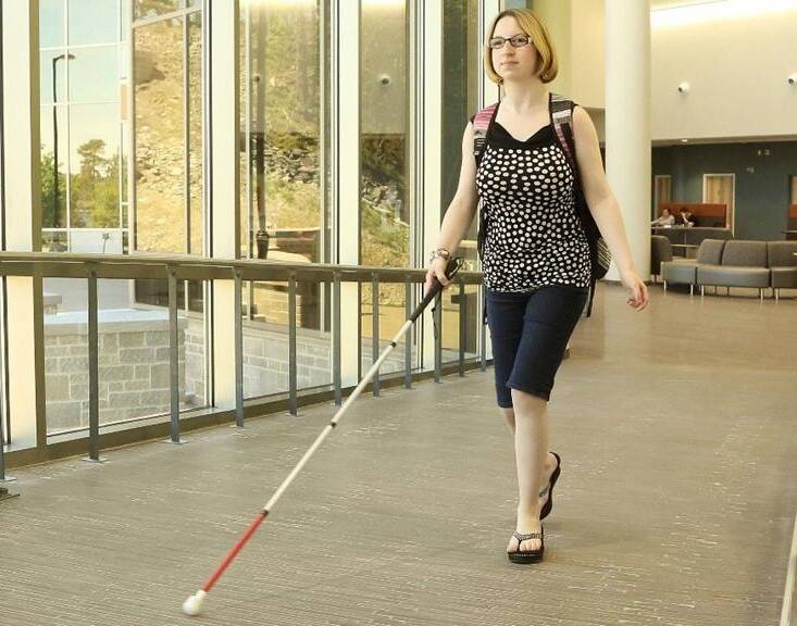 Une femme blanche portant un sac à dos, avançant avec sa canne blanche dans un couloir fenestré.