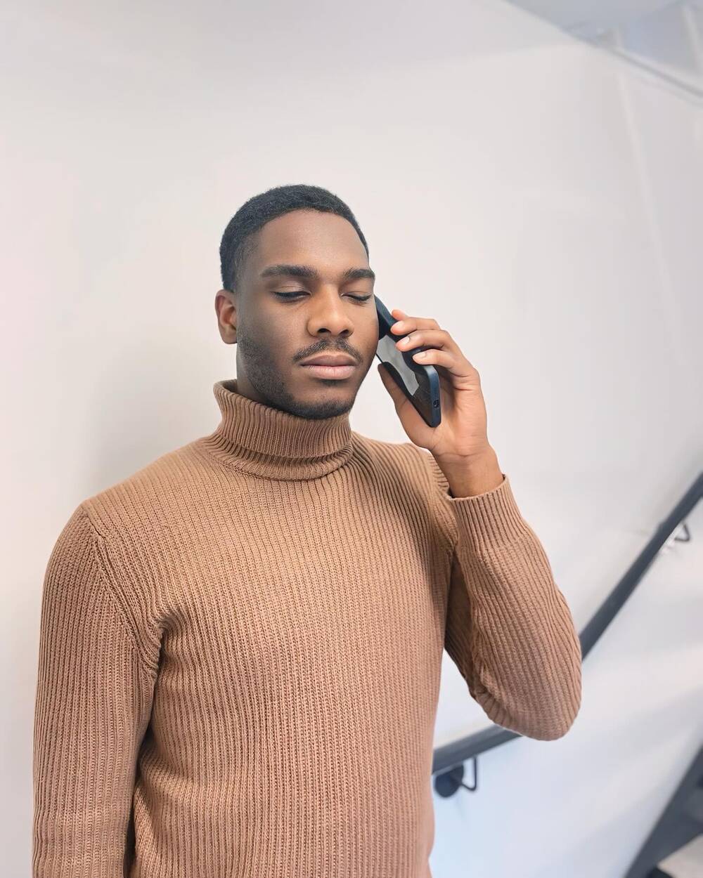 Michel-Antony, un jeune homme noir, parle au téléphone cellulaire. Il porte un chandail à col roulé beige.