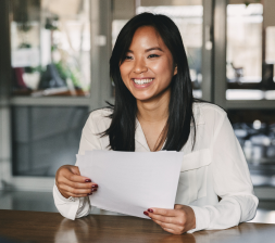 Une jeune femme asiatique vêtue d'un chemisier blanc est assise à un bureau et tient des feuilles de papier. 