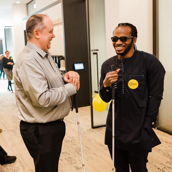 Un jeune homme noir stylé et un homme blanc discutent en souriant . Tous les deux tiennent canne blanche.