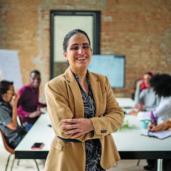 A women with sight loss smiling with her arms crossed. She is standing in front of a group of people sitting around a boardroom table
