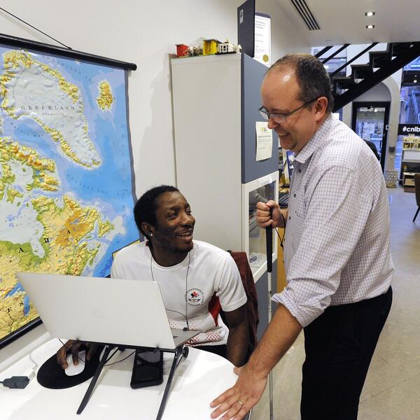 A young man sitting at a computer. Another man holding a white man stands beside him, engaging with the computer
