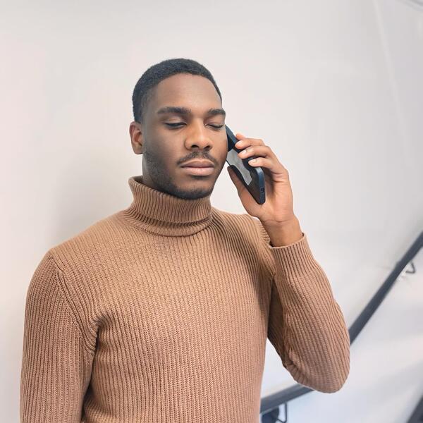 Michel-Antony, un jeune homme noir, parle au téléphone cellulaire. Il porte un chandail à col roulé beige.